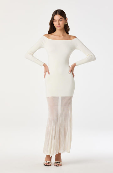 A woman is wearing an off-the-shoulder, long-sleeve, fitted white dress with a semi-sheer pleated skirt that reaches the floor. She has gold hoop earrings and open-toe high-heeled sandals with black and white embellishments.