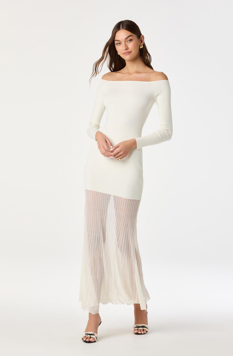 A woman wearing an off-the-shoulder, long-sleeved, fitted white dress with a sheer pleated skirt that extends to the floor. She has dark, wavy hair and is accessorized with gold hoop earrings and black and white heeled sandals.