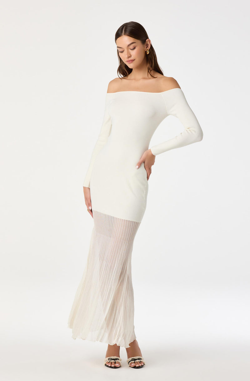 A woman is wearing an off-the-shoulder, long-sleeved white dress that is fitted through the waist and hips. The dress features a pleated, semi-transparent fabric from mid-thigh to the floor, creating a flowing, elegant silhouette. She completes the look with open-toed heels with a bow detail.