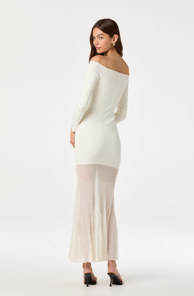 A woman is wearing an off-the-shoulder long-sleeve white dress that fits snugly around her torso and hips. The dress features a pleated, semi-transparent, light beige or blush-colored fabric at the lower part, which extends into wide-leg pants. She is also wearing black high-heeled shoes with ankle straps and gold hoop earrings.
