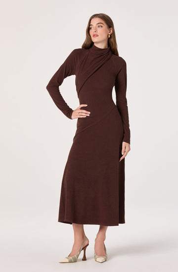 The woman is wearing a long, dark brown dress with a high neckline and long sleeves. The dress has a fitted waist and a slightly flared hem that reaches just above her ankles. She is also wearing a pair of beige and cream patterned high-heeled shoes.