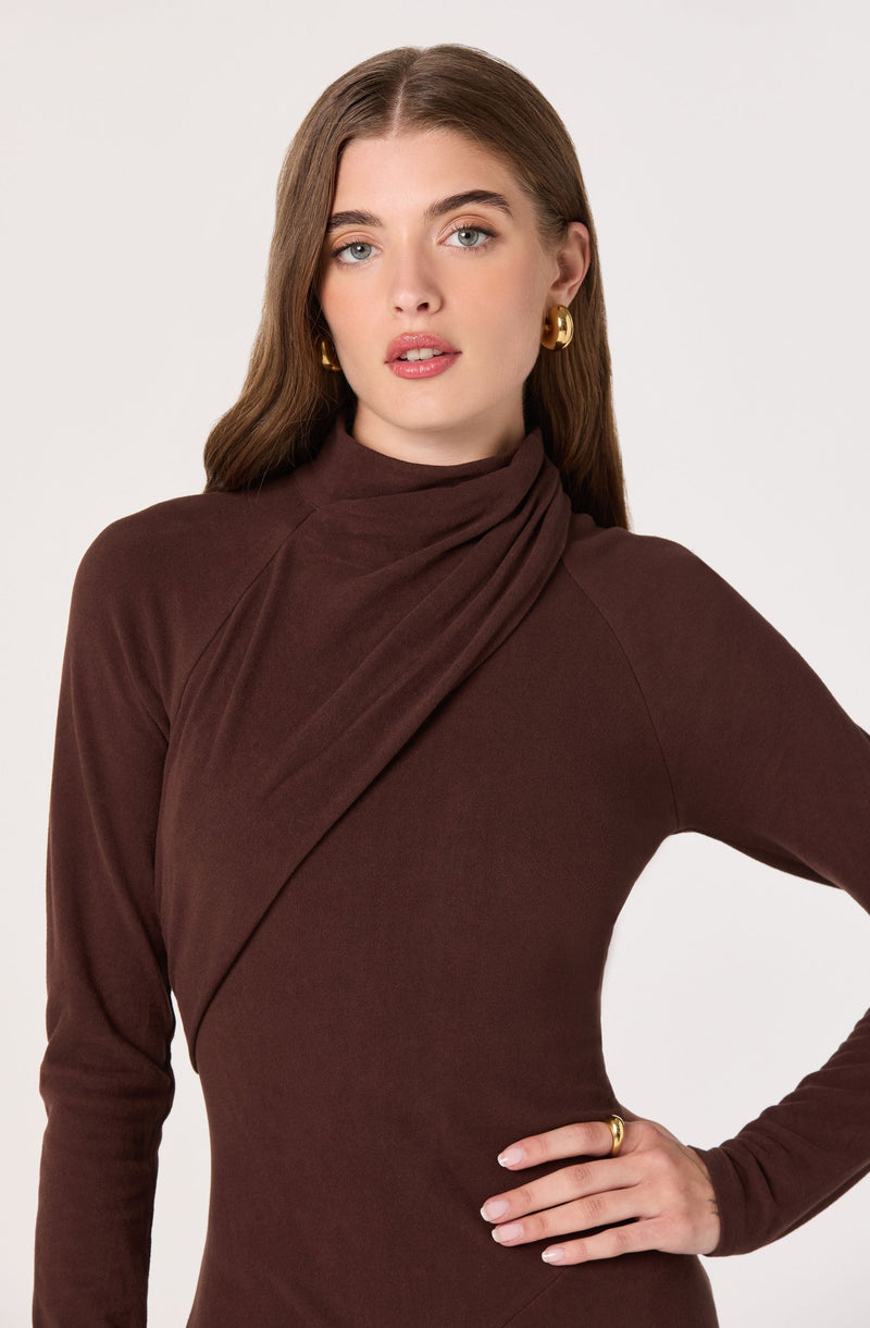 A woman is wearing a fitted, long-sleeved, high-neck dress in a deep brown color. The dress has a draped detail at the shoulder. She accessorizes with large gold hoop earrings and a gold ring on her right hand.