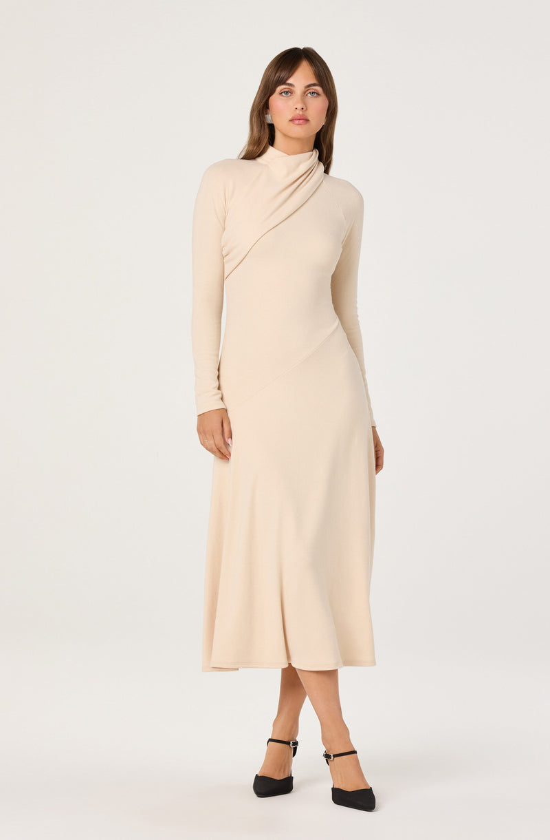 A woman is wearing a long, cream-colored dress with a high neckline and long sleeves. The dress features a subtle drape detail across the chest and a flowy, ankle-length skirt. She is also wearing black heels with ankle straps.
