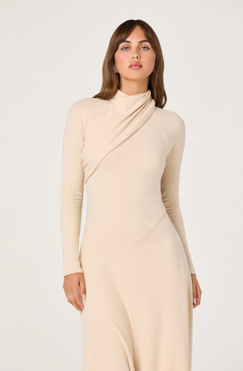 A woman is wearing a long, elegant, cream-colored dress with a high neckline and long sleeves. The dress features soft draping details on the shoulder and subtle seams that create a fitted silhouette.
