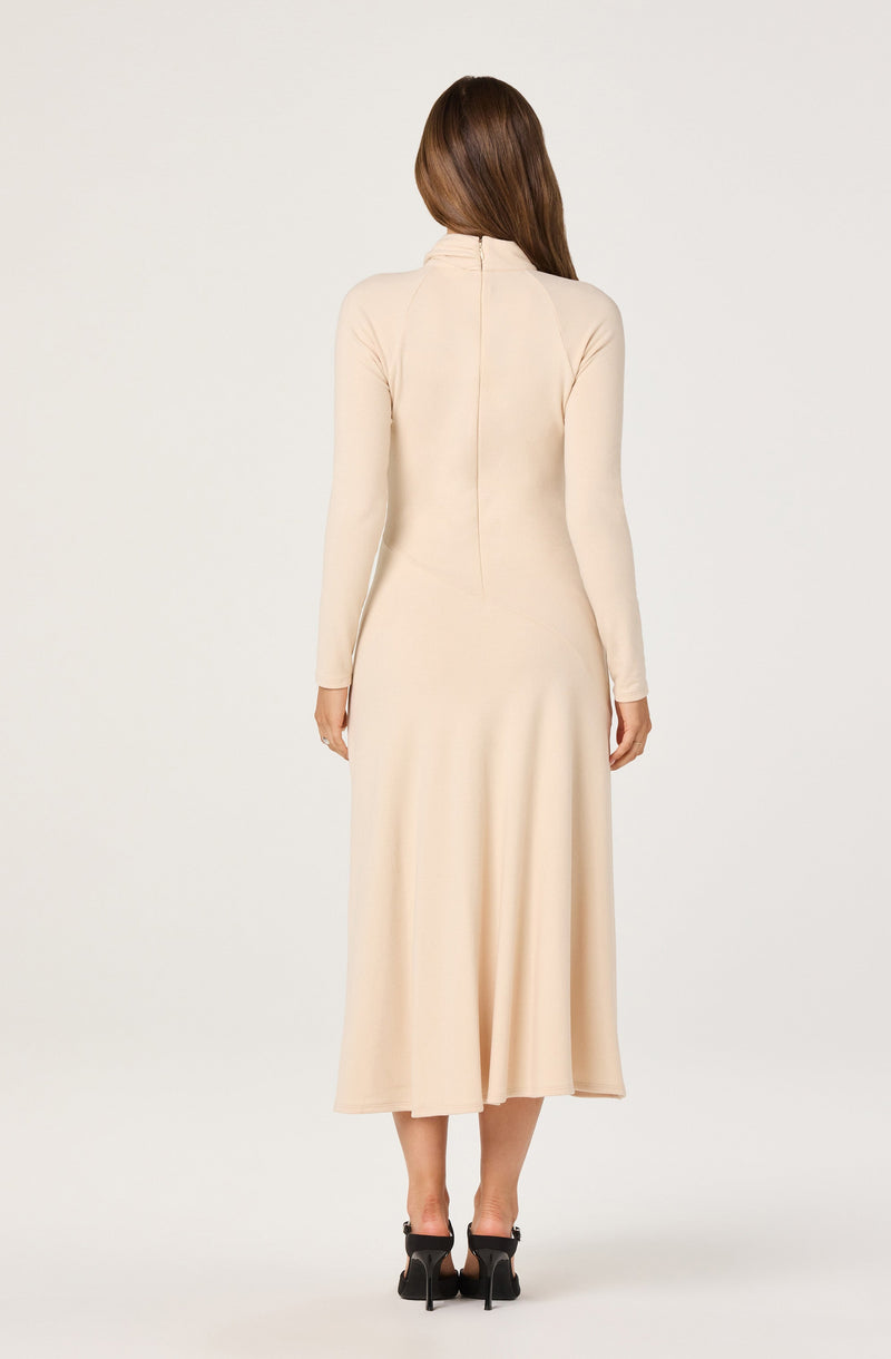 A woman is wearing a long, beige dress with a high collar and a zipper running down the back. The dress is fitted at the top and flows loosely down to the hem, which falls just above her ankles. She is also wearing black high-heeled shoes.