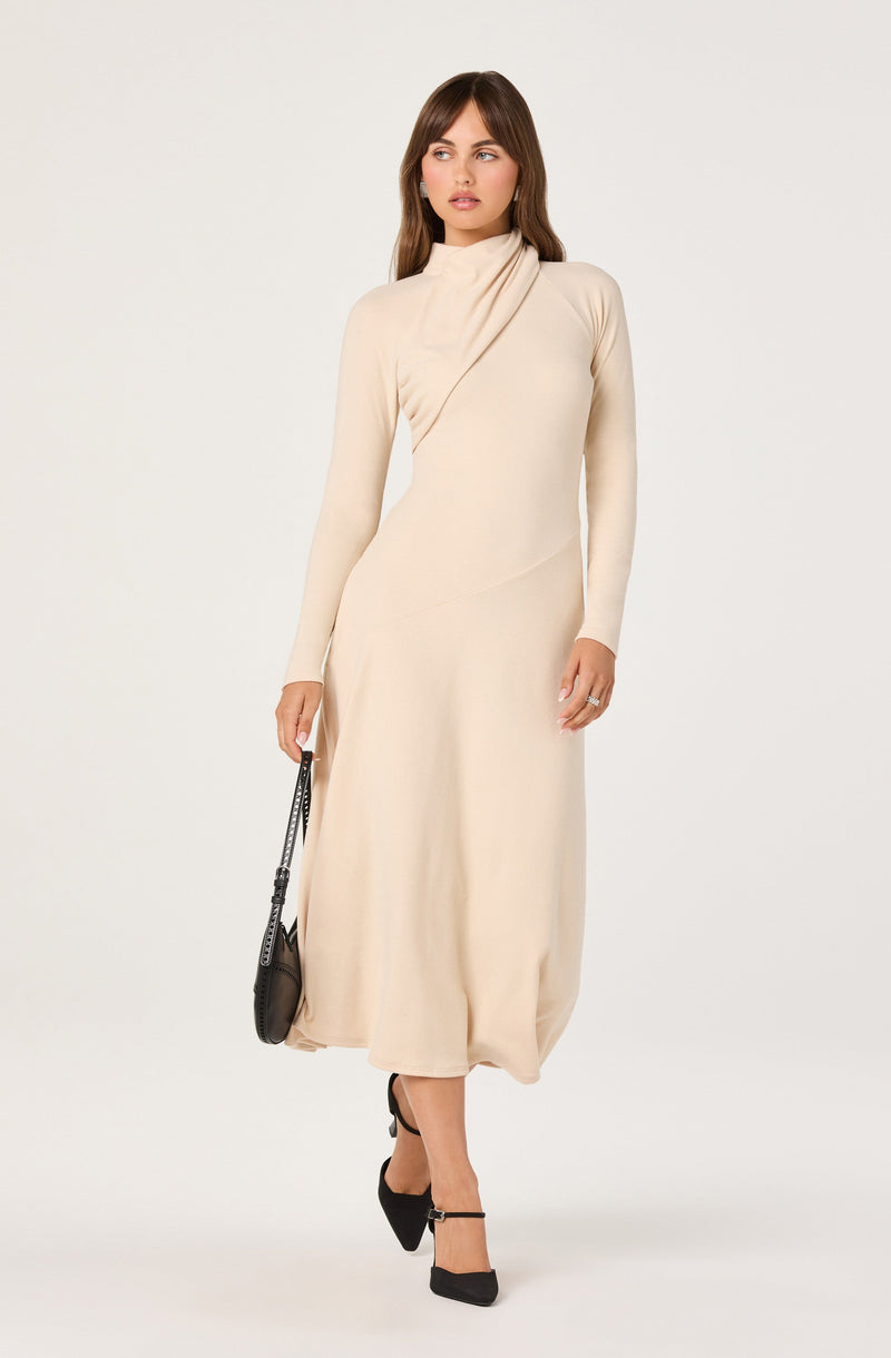 A woman is wearing a long, cream-colored dress with long sleeves and a draped collar that extends over the shoulder. She is holding a small black handbag with a shoulder strap in her right hand. She is also wearing black high-heeled shoes with ankle straps.