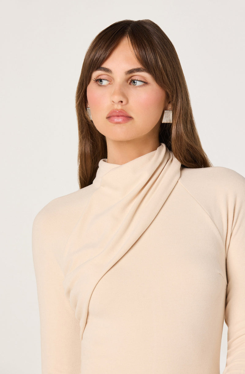 A woman is wearing a high-neck, long-sleeved, cream-colored top with a draped scarf-like detail around the collar area. She has long, straight brown hair and is accessorized with large, rectangular earrings featuring a pattern of vertical lines.