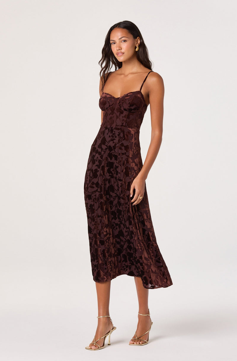 The woman is wearing a sleeveless, dark burgundy dress with thin spaghetti straps. The dress features a fitted bodice with a sweetheart neckline and a floral velvet pattern throughout. It has a midi length, ending below the knees, with a slightly flared hem. She is also wearing gold hoop earrings, a gold ring, and gold high-heeled sandals with thin straps.