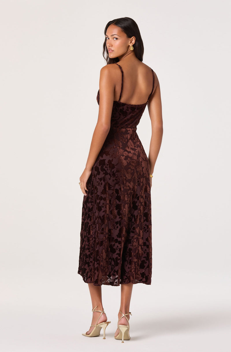 A woman is wearing a sleeveless, ankle-length dress with a fitted bodice and thin spaghetti straps. The dress has a rich, dark reddish-brown color with a floral pattern made of textured floral velvet fabric. She is accessorized with gold hoop earrings, a gold bracelet, and beige strappy high-heeled sandals.