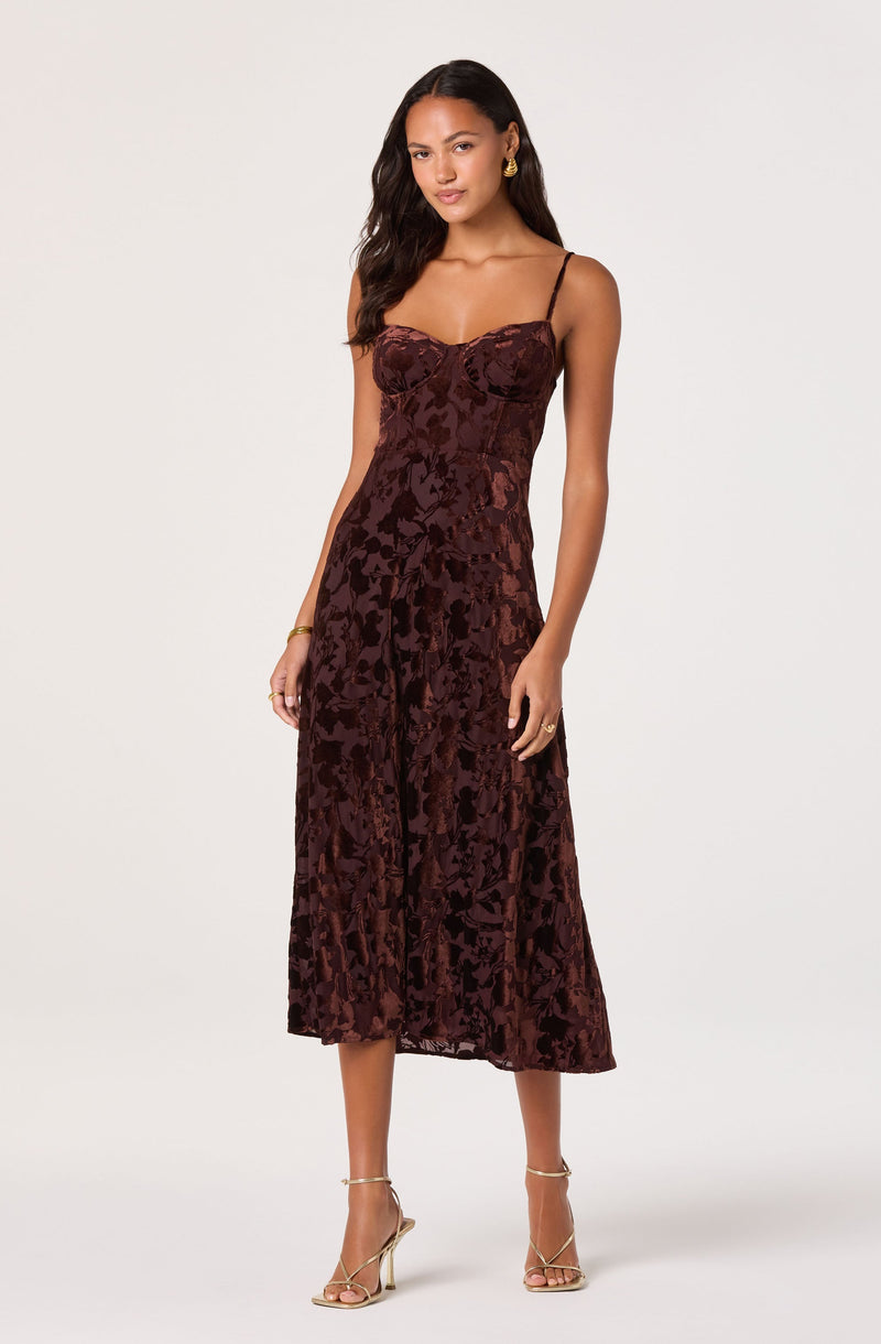 A woman is wearing a sleeveless, dark burgundy dress with thin spaghetti straps. The dress has a fitted bodice and a flowy, A-line skirt that falls to mid-calf length. The fabric features a textured, velvet floral pattern. She is accessorized with gold hoop earrings, a gold bracelet, and rings, and she is wearing strappy high-heeled sandals.