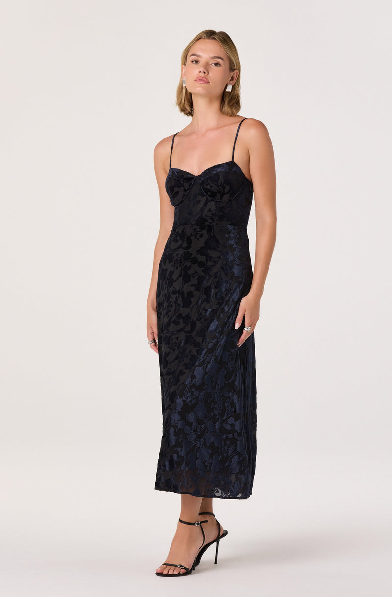 A woman is wearing a long, elegant black dress with a floral pattern. The dress has thin spaghetti straps and a fitted bodice with a bow detail at the center. She is also wearing strappy black high-heeled sandals.