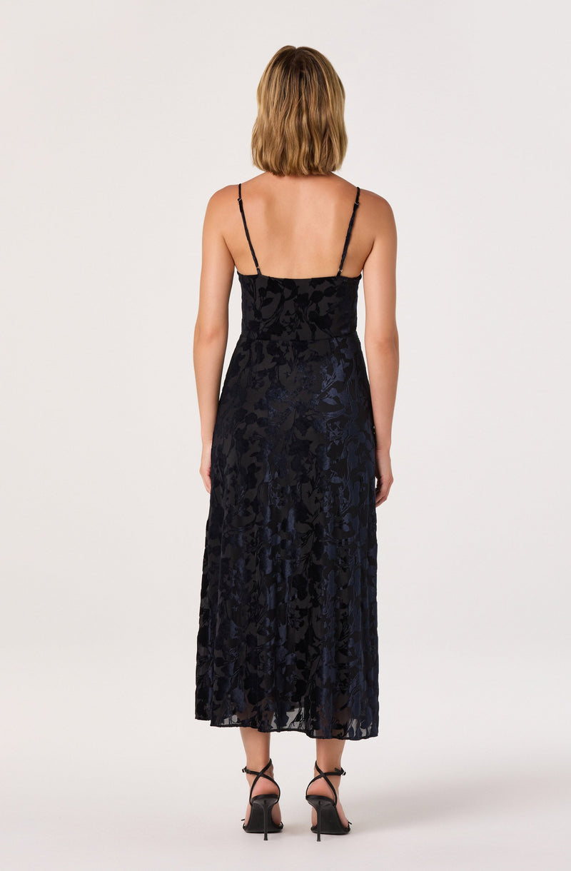 The dress is a sleeveless, full-length gown with thin spaghetti straps. It features a dark, velvety floral pattern throughout the fabric. The dress has a fitted waist and flows down to the hem, which reaches just above the ankles. The fabric appears to have a subtle sheen, adding a touch of elegance to the overall look. The model is wearing black strappy high-heeled sandals.
