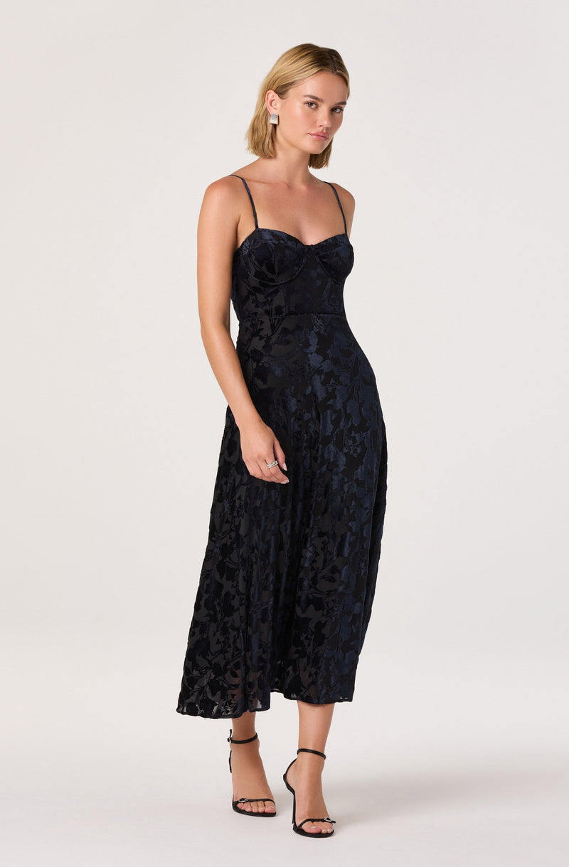 A woman is wearing a black satin dress with thin spaghetti straps. The dress features a fitted bodice with a subtle sweetheart neckline and a floral pattern in a slightly shiny fabric. The skirt is long and flowing, reaching mid-calf length, with a soft, airy appearance. She is also wearing black high-heeled sandals with ankle straps.