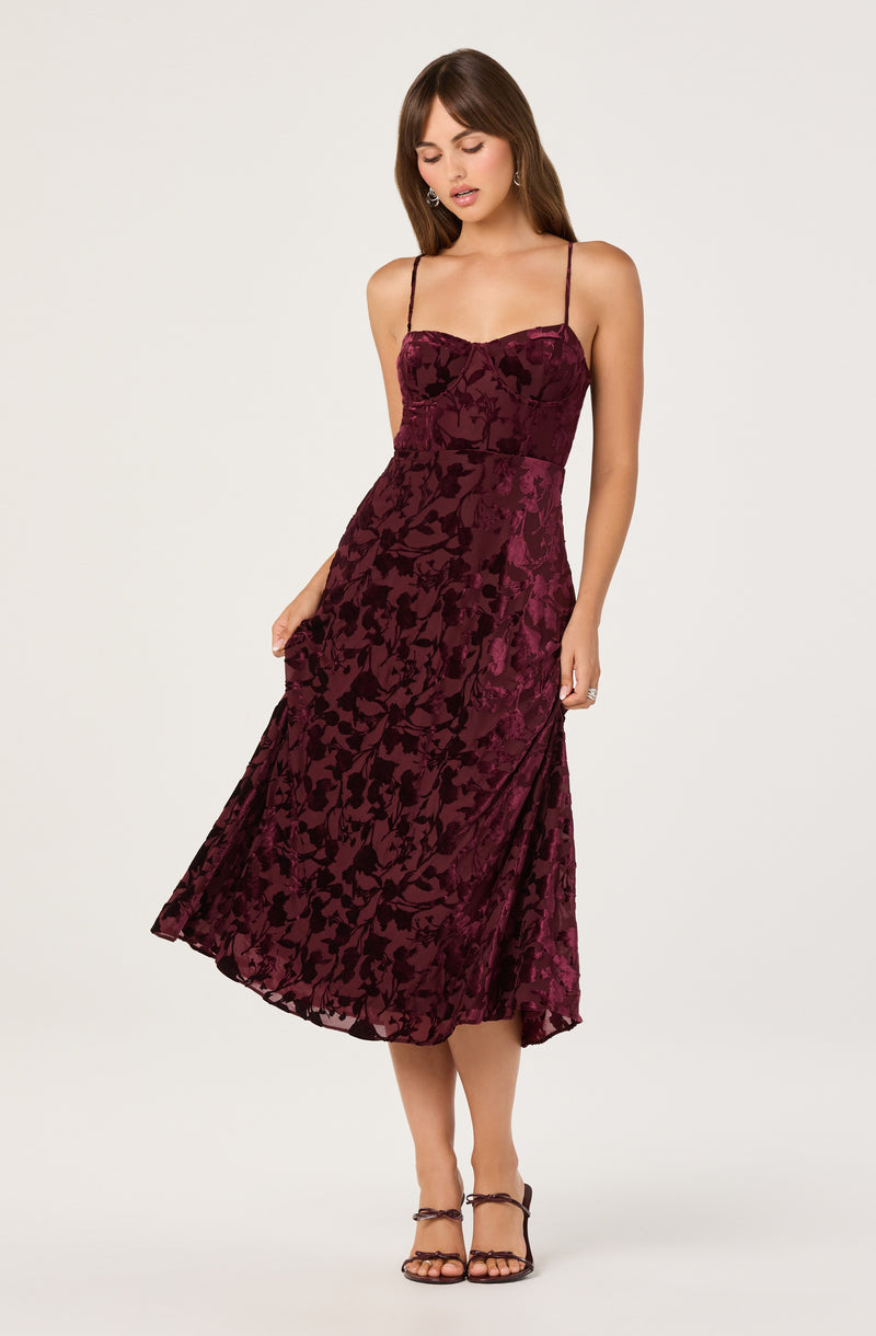 A woman is wearing a sleeveless, spaghetti strap dress with a fitted bodice and a full, mid-length skirt. The dress features a burgundy color with a subtle floral pattern and a slight sheen to the fabric. She is also wearing dark-colored high-heeled sandals.
