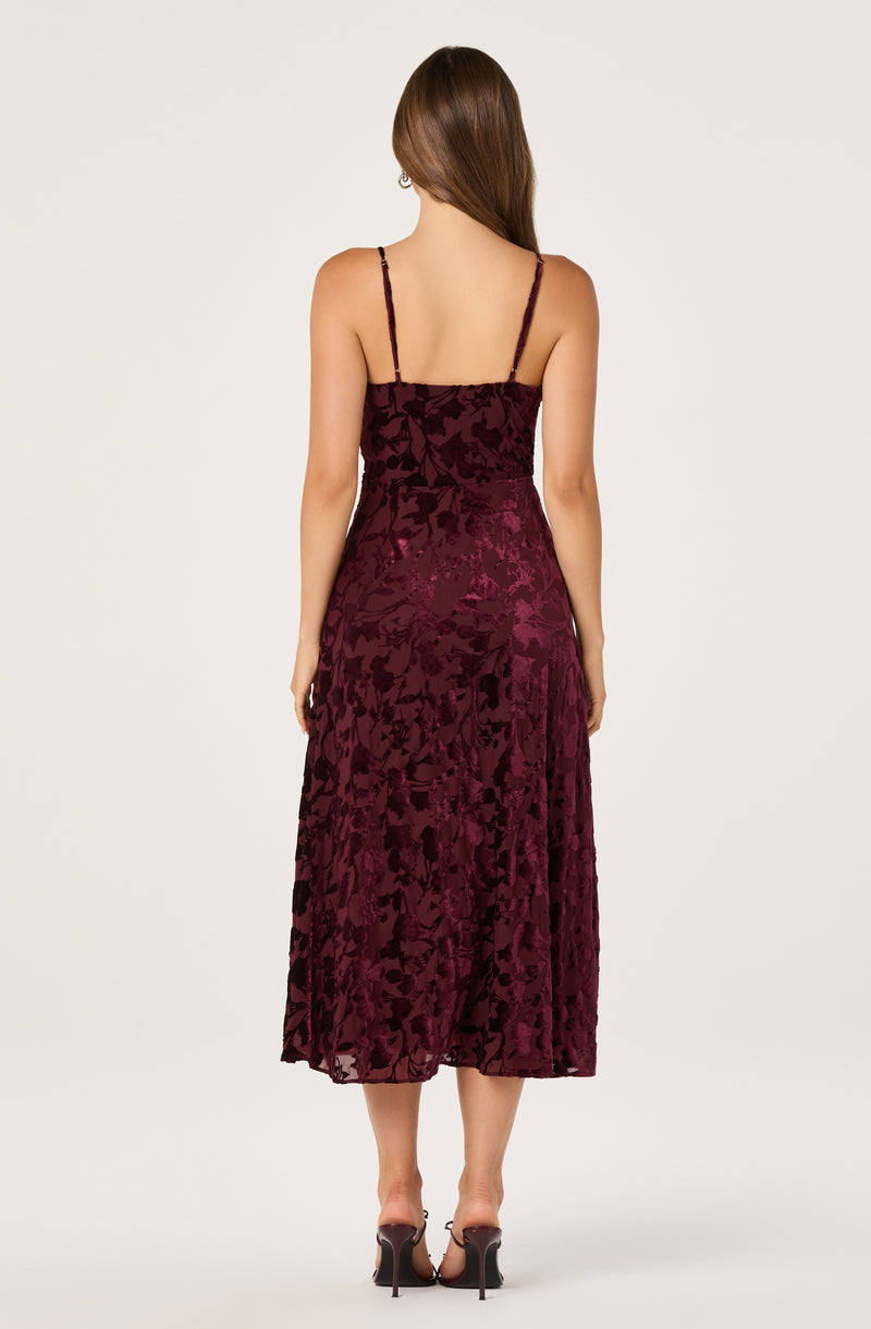 A woman is wearing a sleeveless, burgundy velvet dress with a floral pattern. The dress has thin spaghetti straps and a fitted bodice, flowing into a slightly flared skirt that reaches mid-calf. She is also wearing black high-heeled shoes.