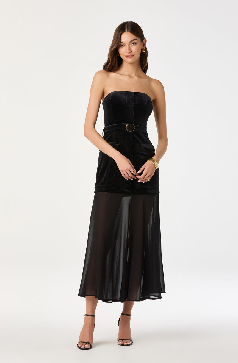 A woman is wearing a strapless black dress with a fitted velvet bodice and a belt with a circular buckle at the waist. The dress has a sheer, flowing black skirt that reaches mid-calf. She is also wearing black strappy high-heeled sandals, gold hoop earrings, and a gold bracelet on her left wrist.