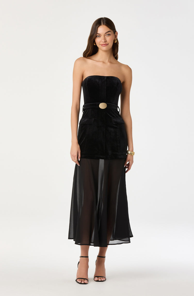 A woman is wearing a strapless black dress with a fitted velvet bodice and a large oval buckle at the waist. She has on sheer, black, wide-leg pants underneath, which are see-through and reach her ankles. She has accessorized with gold hoop earrings, a gold bracelet, and black high-heeled sandals.