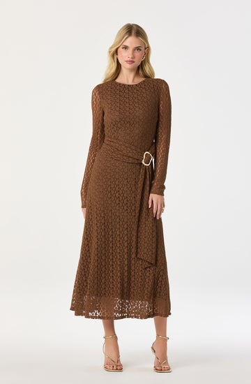 A woman is wearing a matching brown lace dress with long sleeves and a round neckline. The dress features a textured pattern and is fitted at the waist with a belt that has a large gold circular buckle. The skirt is ankle-length with a slightly scalloped hem. She is also wearing open-toe high-heeled sandals with thin straps.