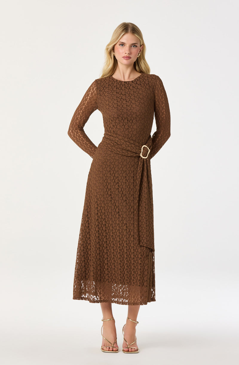 A woman is wearing a long brown dress with a textured, lace-like pattern. The dress has long sleeves, a round neckline, and a waistline accentuated with a matching fabric belt that features a large, oval-shaped gold buckle. She is also wearing high-heeled sandals with thin straps around her ankles.