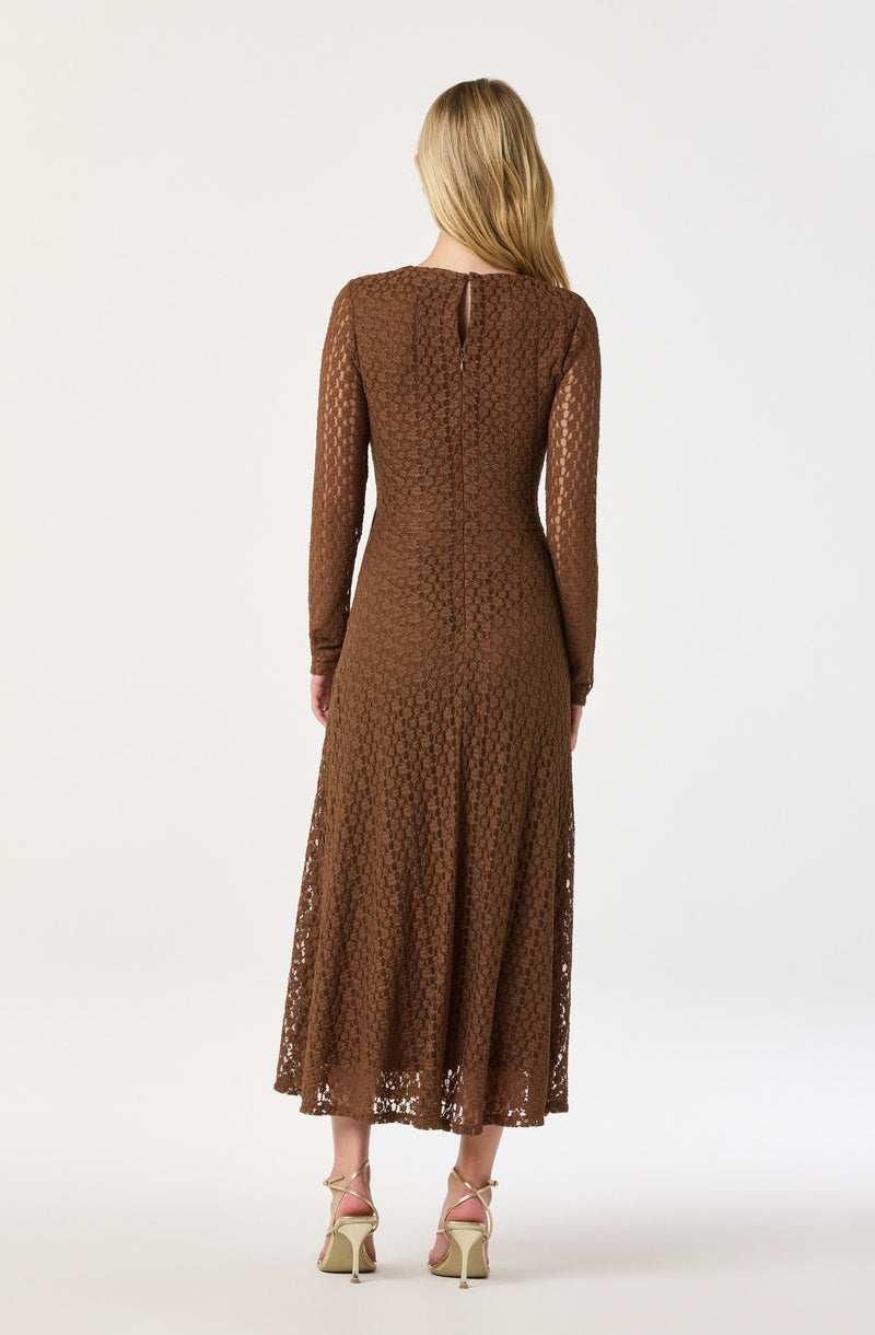 A woman is wearing a long, brown lace dress with long sleeves and a fitted waist. The dress has a sheer, patterned fabric and a keyhole opening at the back with a small button closure at the neckline. She is also wearing beige high-heeled sandals.