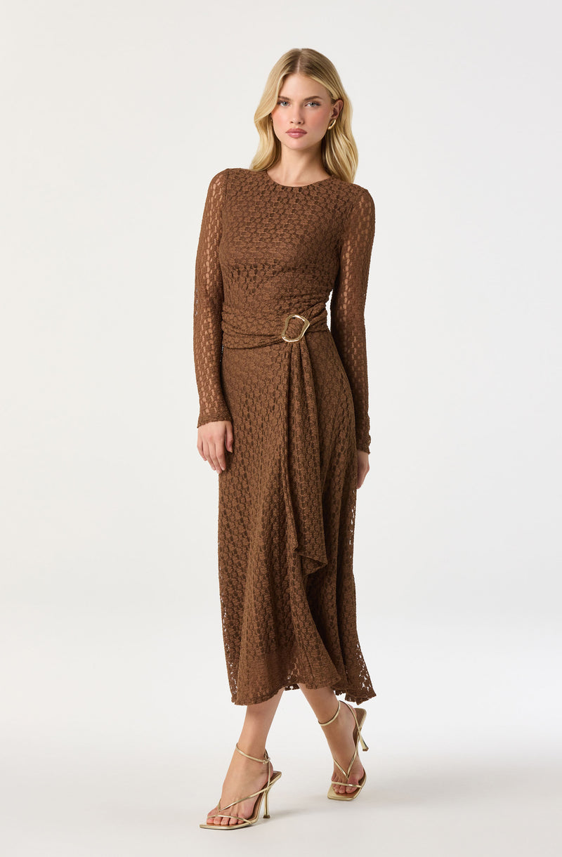A woman is wearing a brown, long-sleeved, ankle-length dress with a textured, patterned fabric. The dress features a round neckline and a matching fabric belt with a ring detail at the waist, creating a gathered effect on the skirt. She is also wearing beige high-heeled sandals with thin straps.