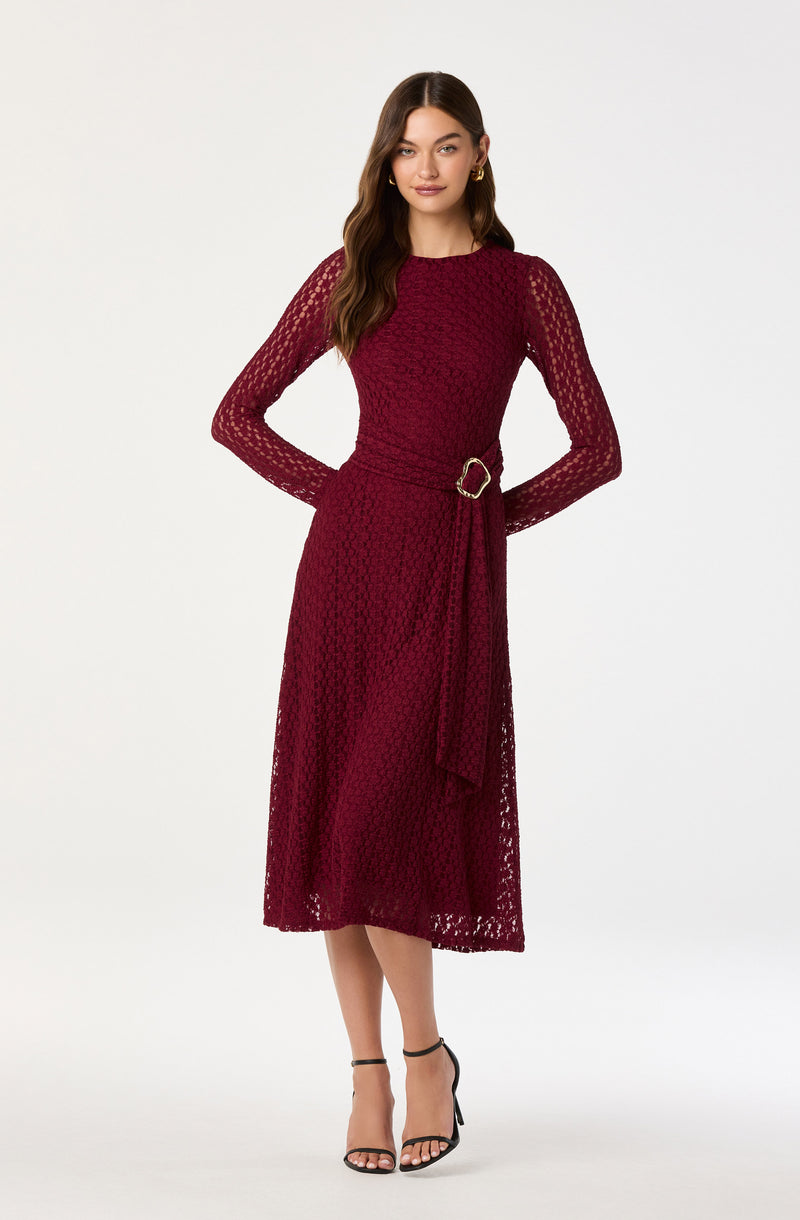 A woman is wearing a long, burgundy lace dress with long sheer sleeves and a round neckline. The dress features a belted waist with a decorative gold buckle and a skirt that falls below the knees. She has paired the outfit with black strappy high-heeled sandals.