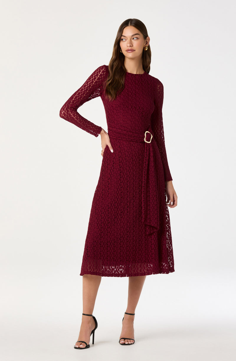 A woman is wearing a long, burgundy lace dress with long sleeves and a round neckline. The dress features a belt with a gold buckle at the waist and has a textured, open lace pattern throughout. She is also wearing black high-heeled sandals with ankle straps.