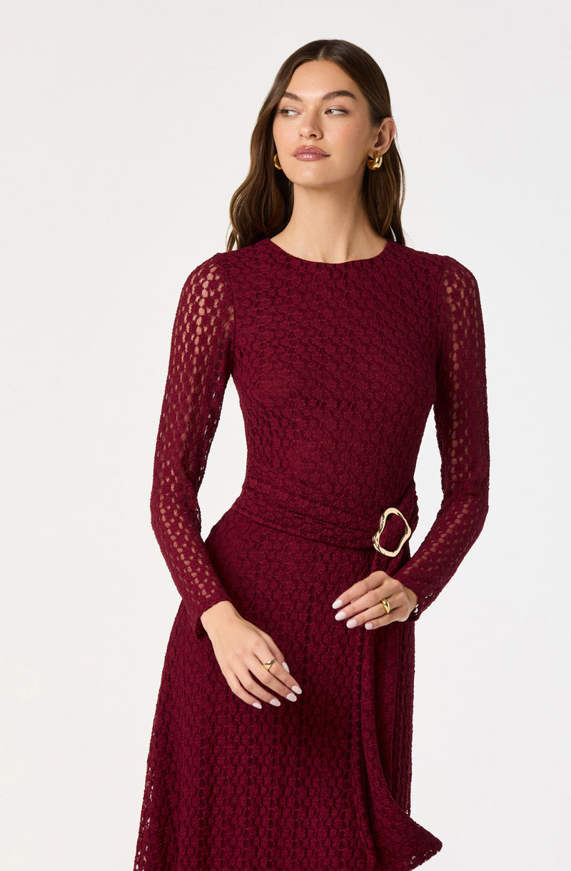 A woman is wearing a fitted, long-sleeved, deep red lace dress with a round neckline. The dress features a textured pattern and a matching belt with a large gold buckle at the waist. She has gold hoop earrings and gold rings on her fingers. Her hair is styled in loose waves, and she has a neutral makeup look.