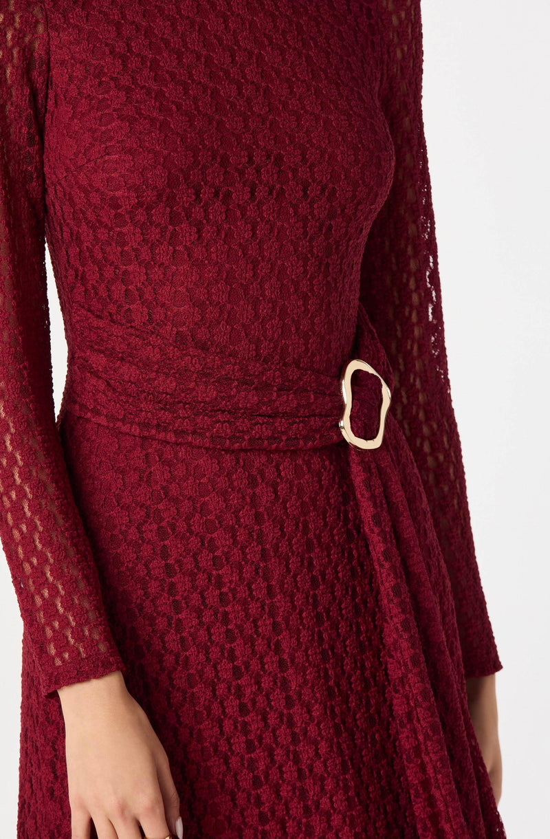 A woman is wearing a long, fitted, deep red dress made of textured lace fabric with a honeycomb pattern. The dress features long sleeves, a high neckline, and a matching belt at the waist with a large, irregularly shaped, gold-colored buckle.