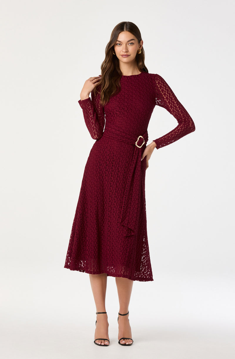 A woman is wearing a long, burgundy lace dress with long sleeves and a round neckline. The dress features a textured floral pattern with a matching belt that has a gold buckle, tying at the waist. She completes her outfit with black strappy high heels.