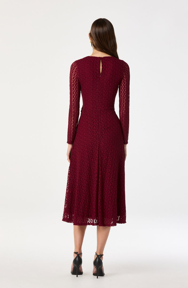 A woman is wearing a long, burgundy lace dress with long sheer lace sleeves. The dress has a fitted waist and a keyhole opening at the back near the neckline. She is also wearing black high-heeled shoes.
