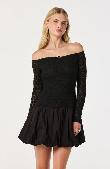 A woman is wearing an off-the-shoulder black lace top with long sleeves and a small bow detail at the neckline. She pairs it with a black, voluminous, bubble-hem skirt made of shiny fabric.