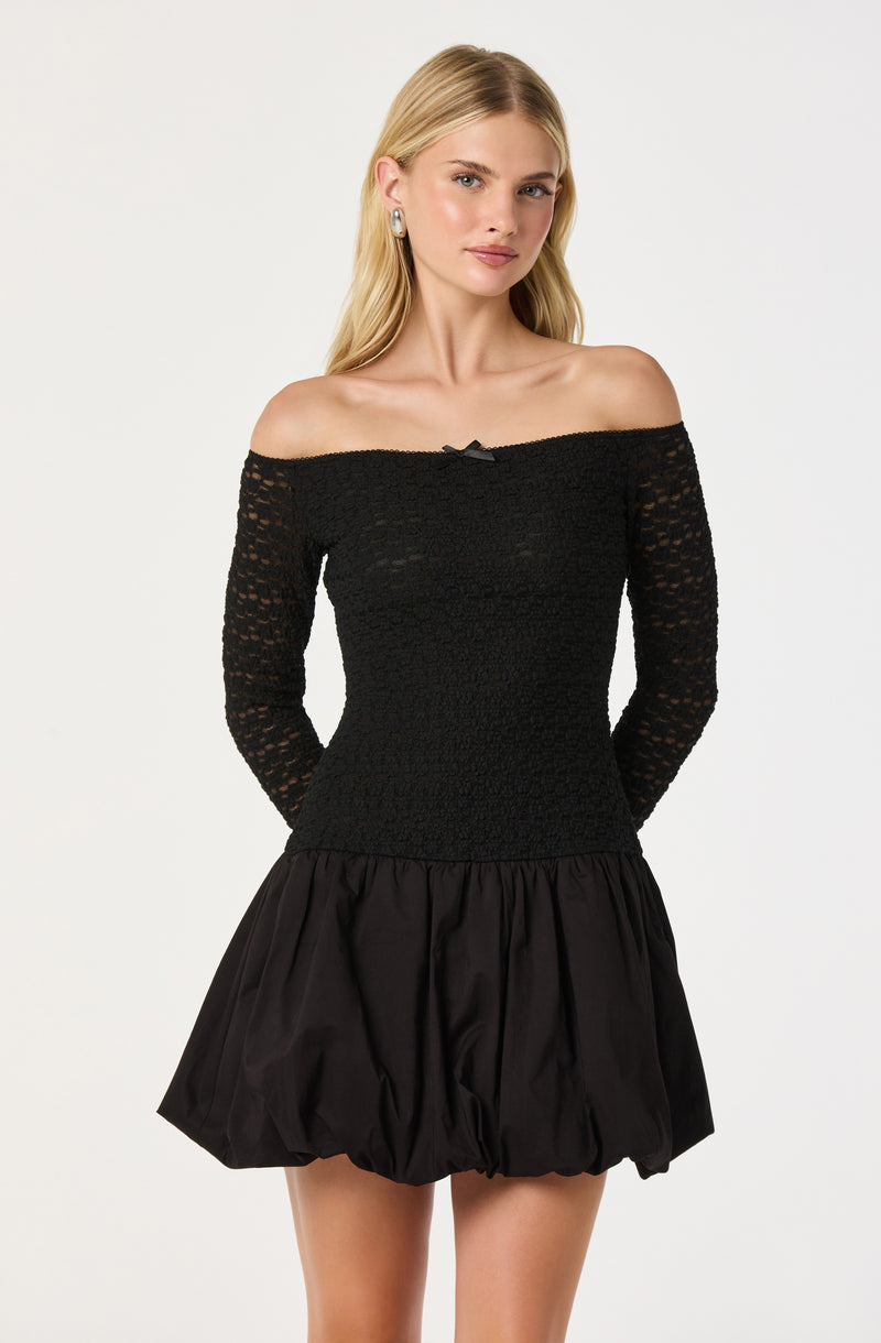 The woman is wearing a black, off-the-shoulder dress. The top part is made of a textured, open-knit fabric with long sleeves, and features a small bow at the center of the neckline. The skirt is a voluminous, gathered style with a shiny, satin-like finish.
