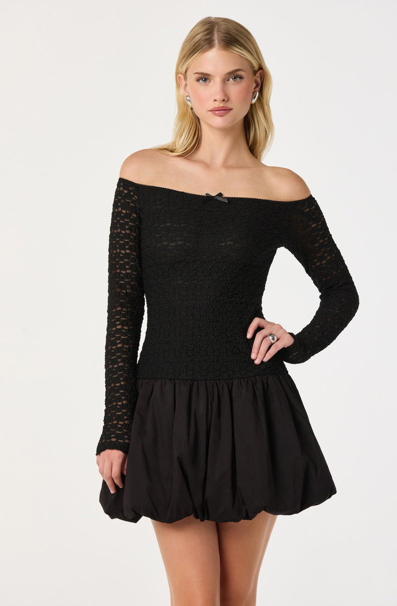 A woman is wearing a black off-the-shoulder long-sleeve top with a textured, open-knit pattern and a small decorative bow at the center of the neckline. She pairs it with a voluminous, bubble-style skirt made of a shiny, slightly gathered fabric.
