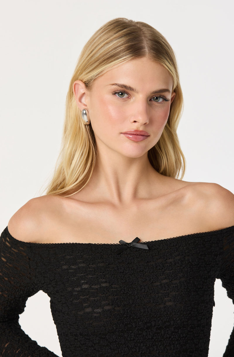 A young woman with fair skin and blonde hair is wearing a black, off-the-shoulder top made of a textured, lace-like fabric. The top has a small black bow in the center of the neckline.