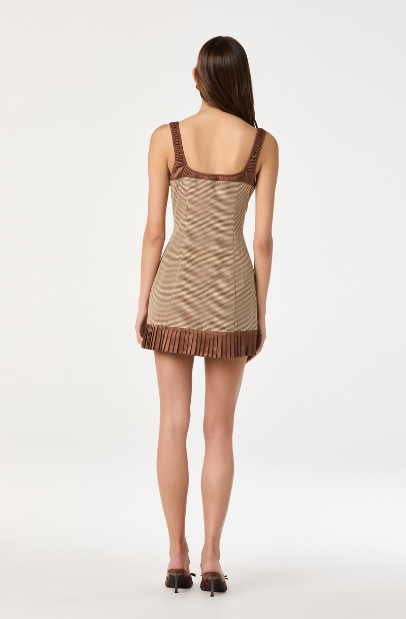 The dress features wide straps with a textured, ruffled trim. The bodice and main body of the dress have a small checkered pattern in neutral tones. The hem of the dress has a pleated, darker brown trim that adds contrast to the lighter fabric.
