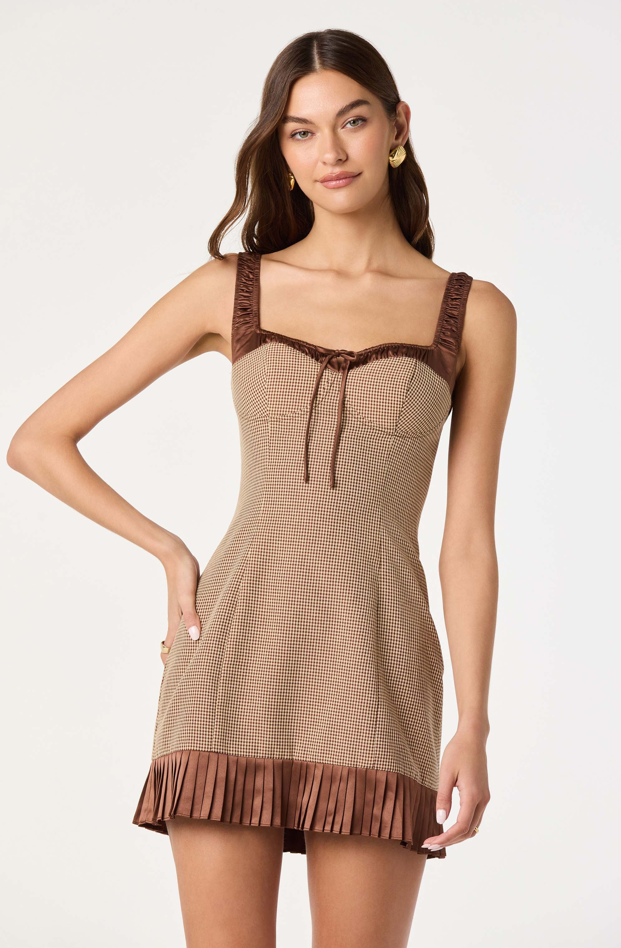 A young woman is wearing a sleeveless dress with wide, gathered straps. The dress has a checkered pattern in beige and brown, with a brown ruffle trim at the neckline accented by a tie at the center front. The hem features a pleated brown fabric that adds texture to the bottom of the dress.