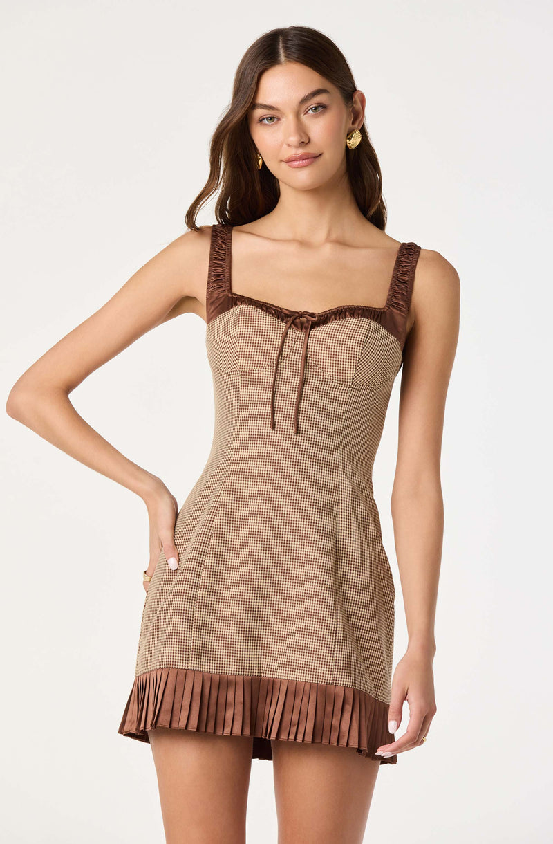 A young woman is wearing a sleeveless dress with wide, gathered straps. The dress has a checkered pattern in beige and brown, with a brown ruffle trim at the neckline accented by a tie at the center front. The hem features a pleated brown fabric that adds texture to the bottom of the dress.