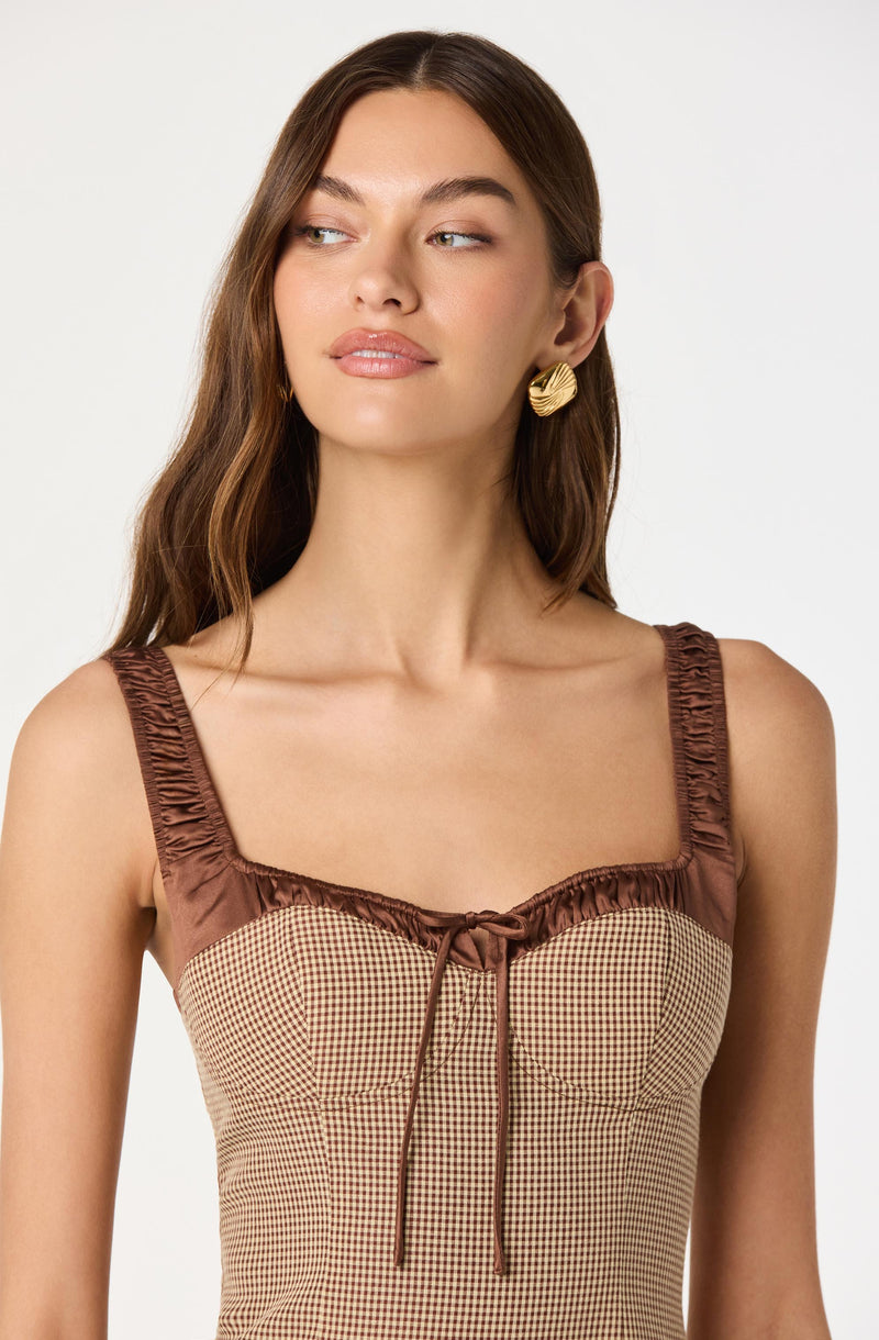 A young woman with long, wavy brown hair is wearing a sleeveless top with ruched straps and a square neckline. The top features a checkered pattern in beige and brown, with a satin-like brown trim and bow detail at the center of the neckline. She is also wearing large, textured gold hoop earrings.