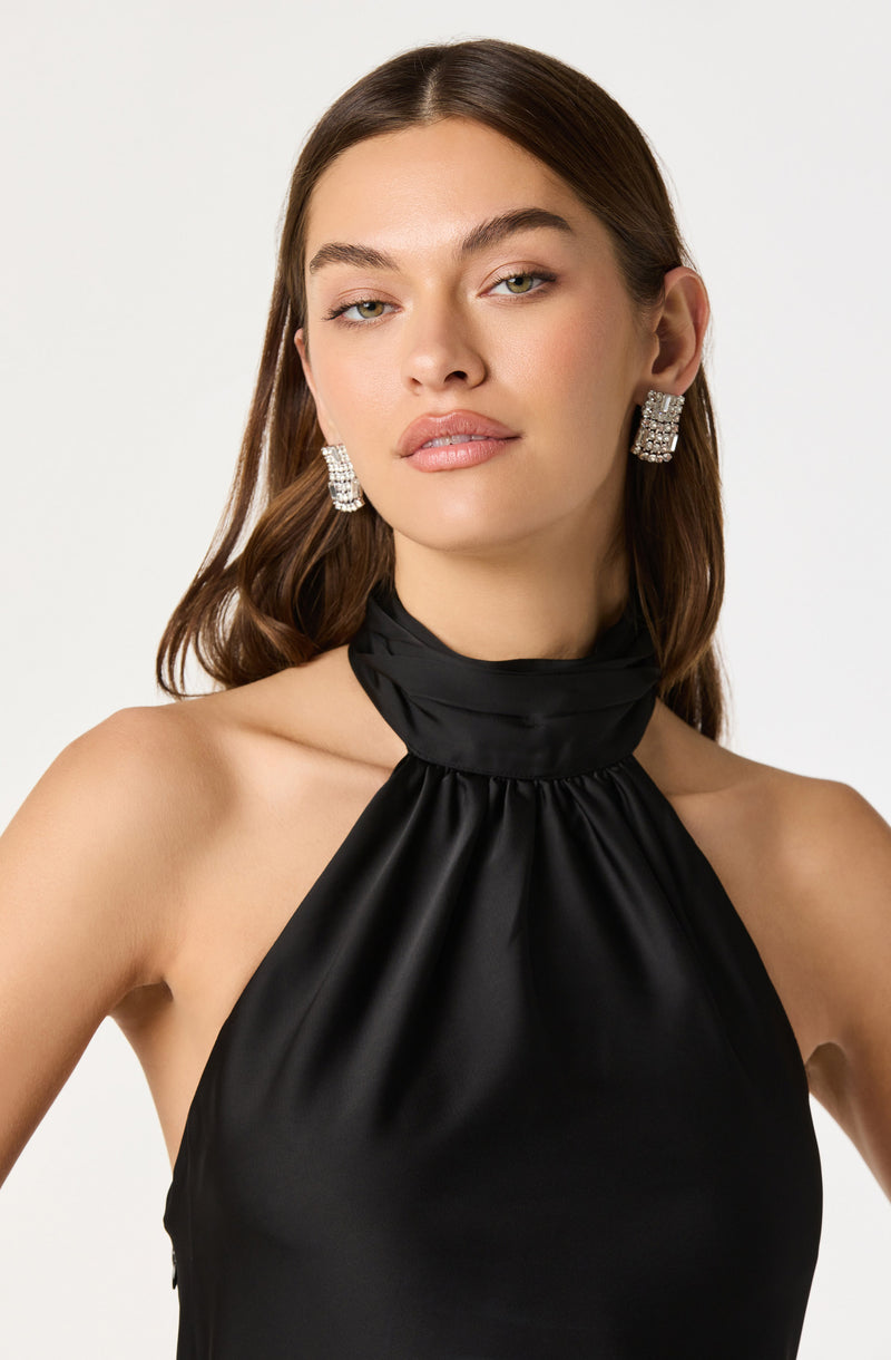 A woman is wearing a sleeveless, high-neck black satin top with gathered fabric at the neckline. She has on large, square, rhinestone-encrusted earrings.
