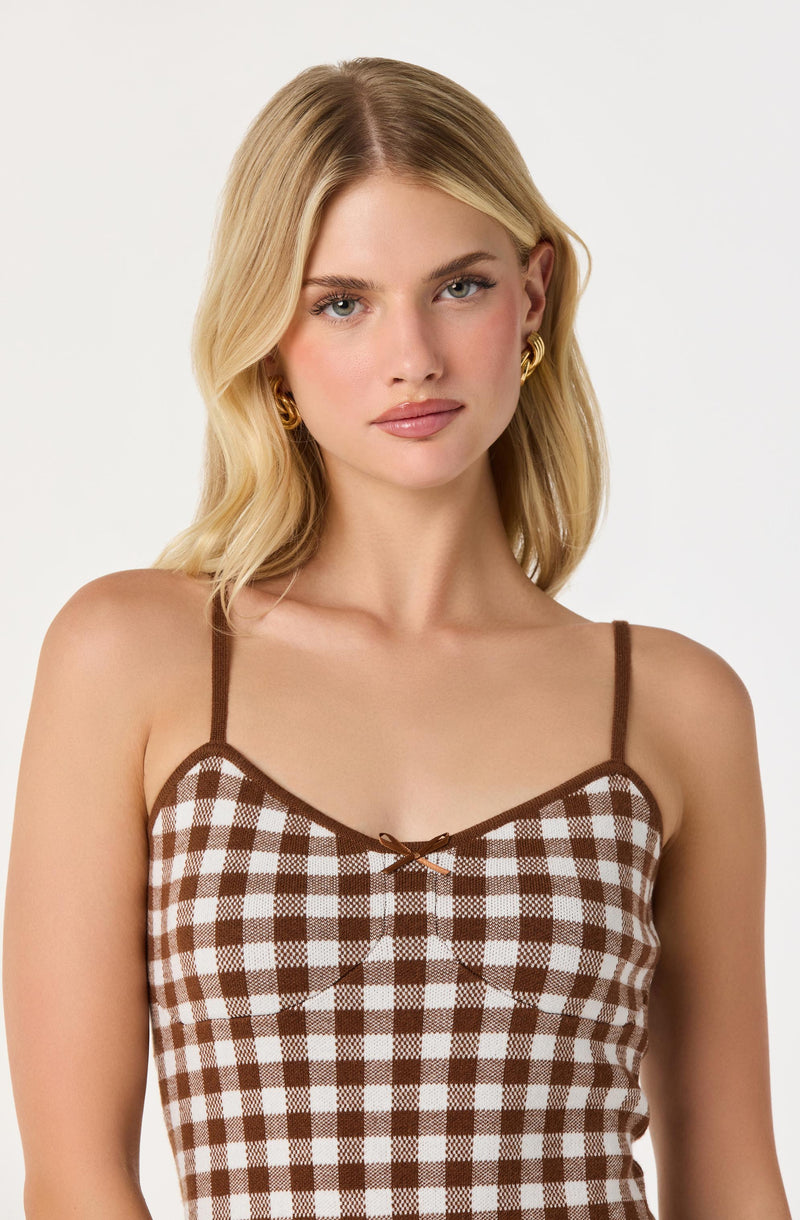 A young woman is wearing a sleeveless, fitted dress with thin spaghetti straps. The dress features a brown and white checkered pattern and has a small decorative brown bow at the center of the neckline. She accessorizes with gold hoop earrings.