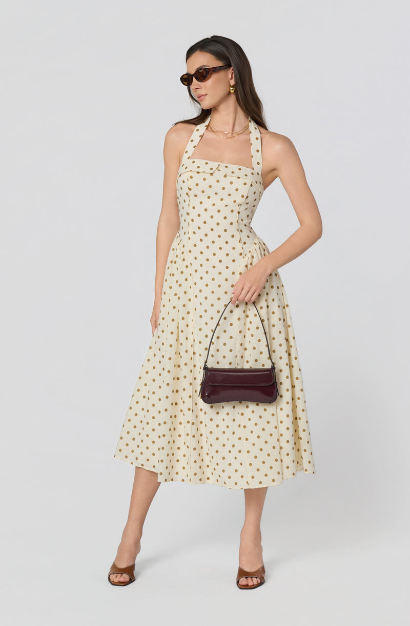 Woman in a cream halter dress with gold polka dots, wearing sunglasses, gold earrings, and brown high heels.
