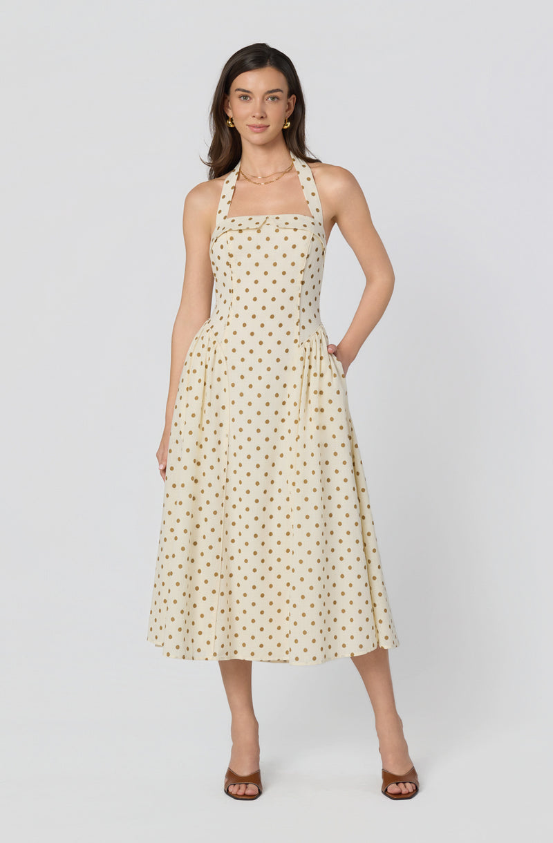 Woman wearing a cream-colored dress with gold polka dots, halter neckline, and side pockets.