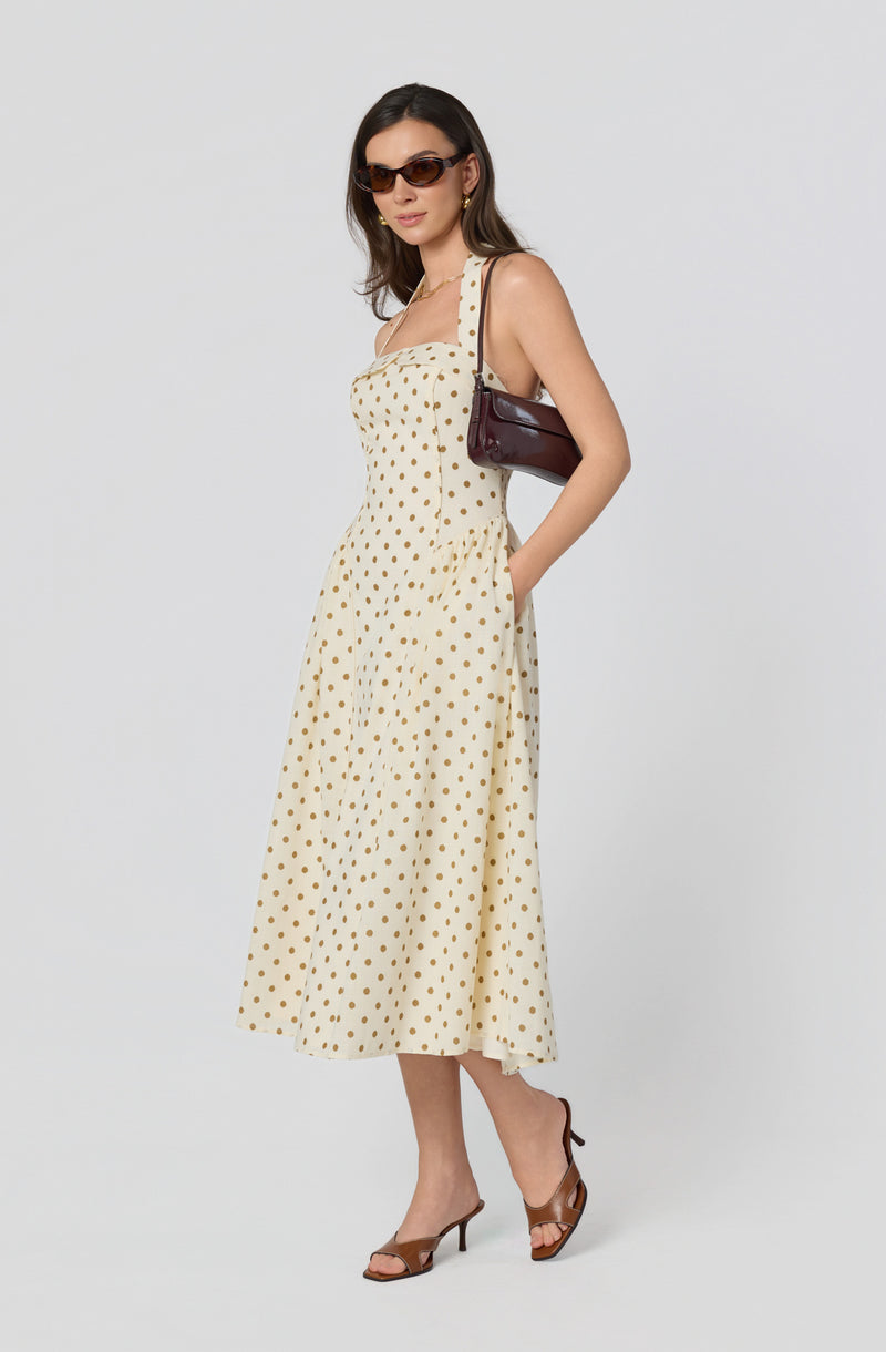 Woman wearing a cream-colored, polka-dot dress with wide straps and a fitted bodice, paired with brown heels, sunglasses, and a dark handbag over her shoulder.
