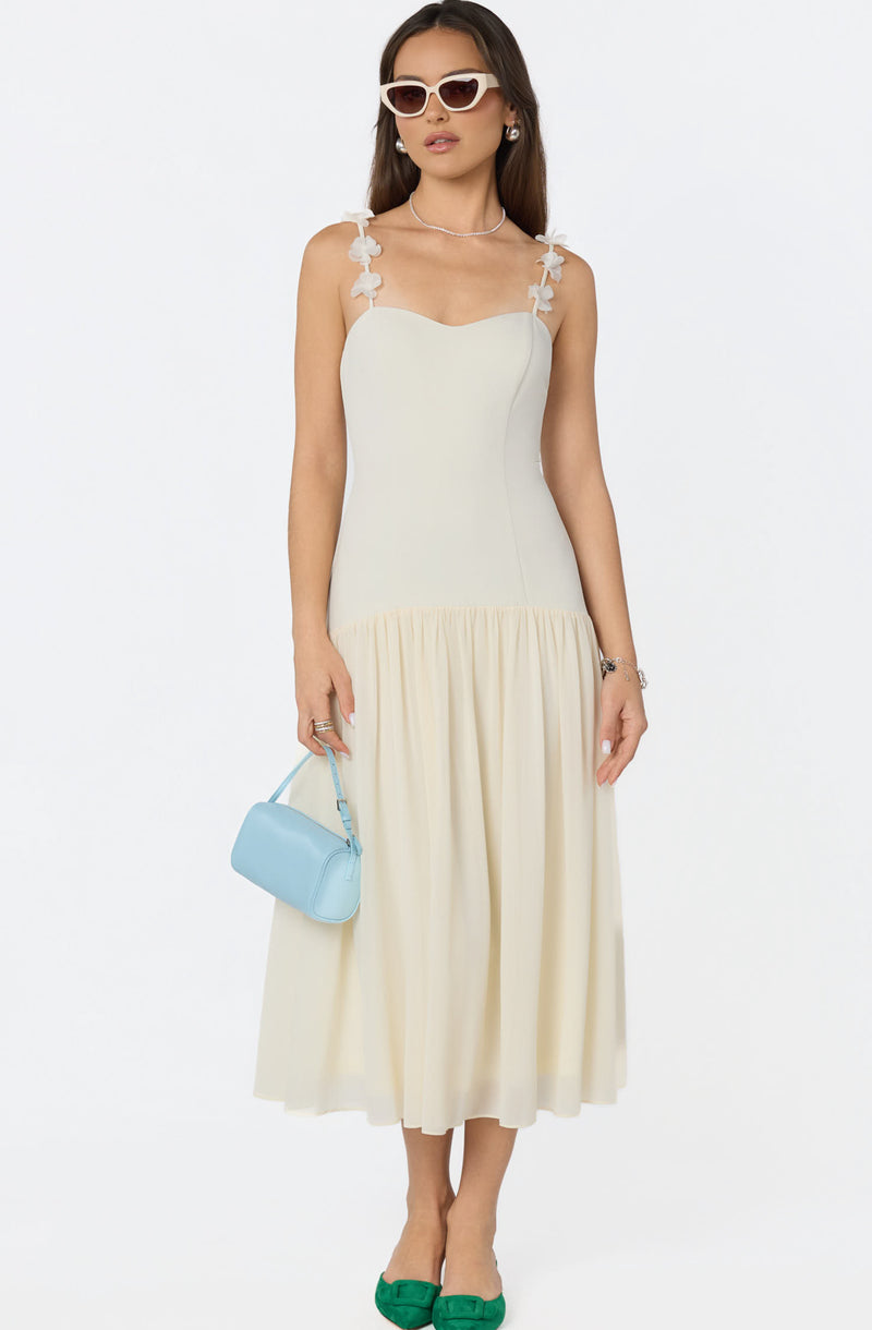 Woman in a white dress with floral shoulder straps, holding a light blue purse, wearing green shoes and white sunglasses.
