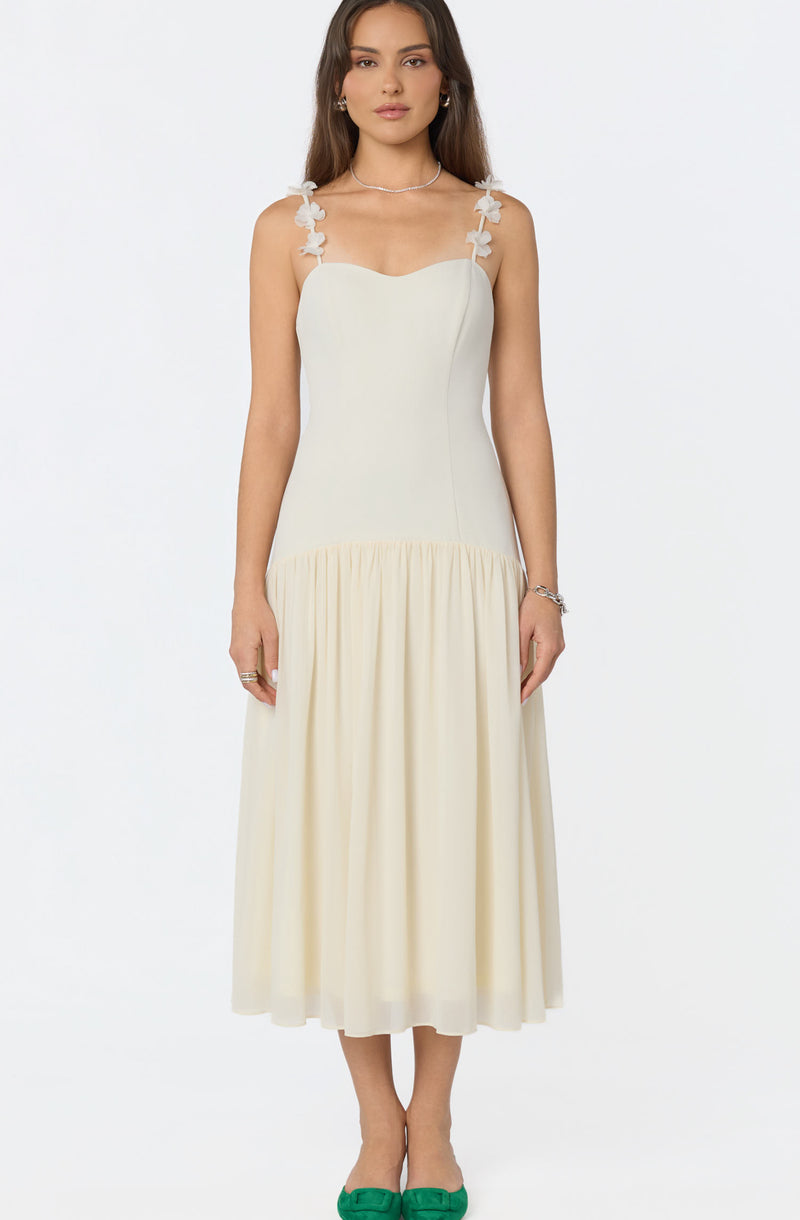 Woman wearing a cream-colored sleeveless dress with floral strap details and a gathered skirt, paired with green shoes.