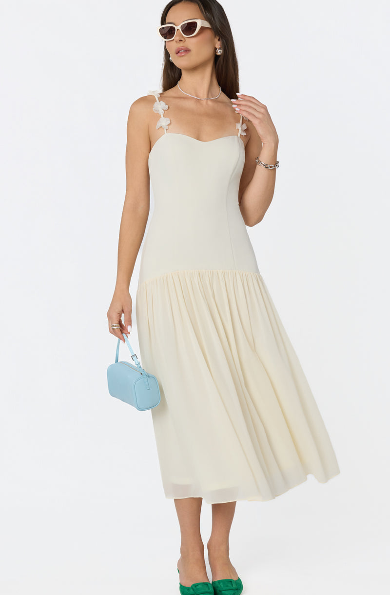 Woman in a cream dress with floral straps, white sunglasses, and green shoes, holding a small blue handbag.