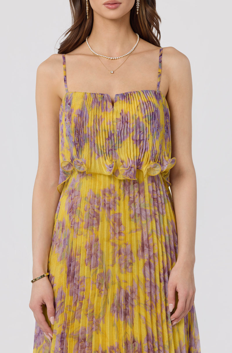 Woman in yellow and purple floral pleated dress with ruffled trim, thin shoulder straps.