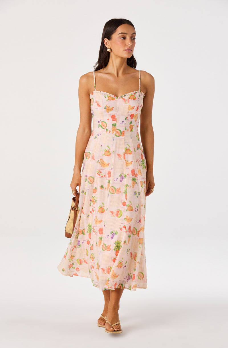 Woman in a pink sundress with a fruit and vegetable print, spaghetti straps, and ruffled neckline.