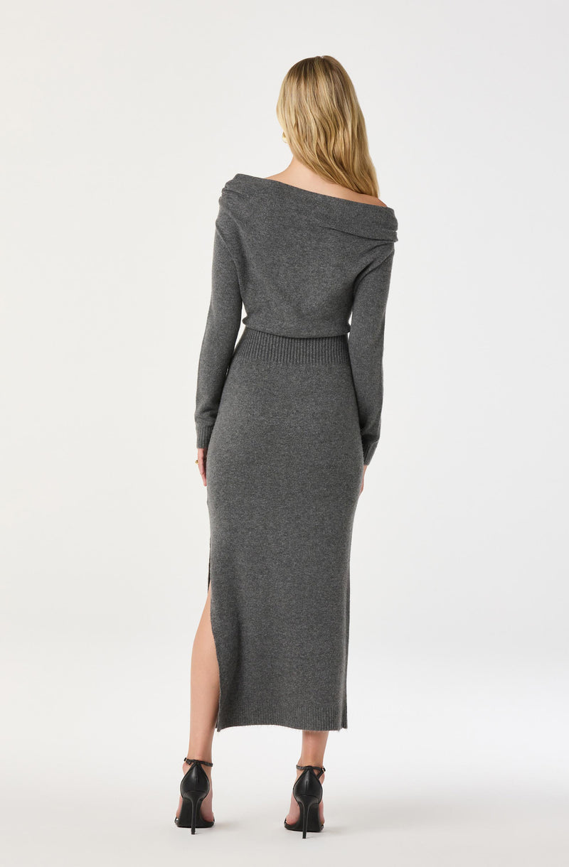 A woman is wearing a long, dark gray knit dress with long sleeves and a wide, off-the-shoulder neckline. The dress has a side slit that reaches up to her knee, revealing her legs. She is also wearing black high-heeled shoes with ankle straps.